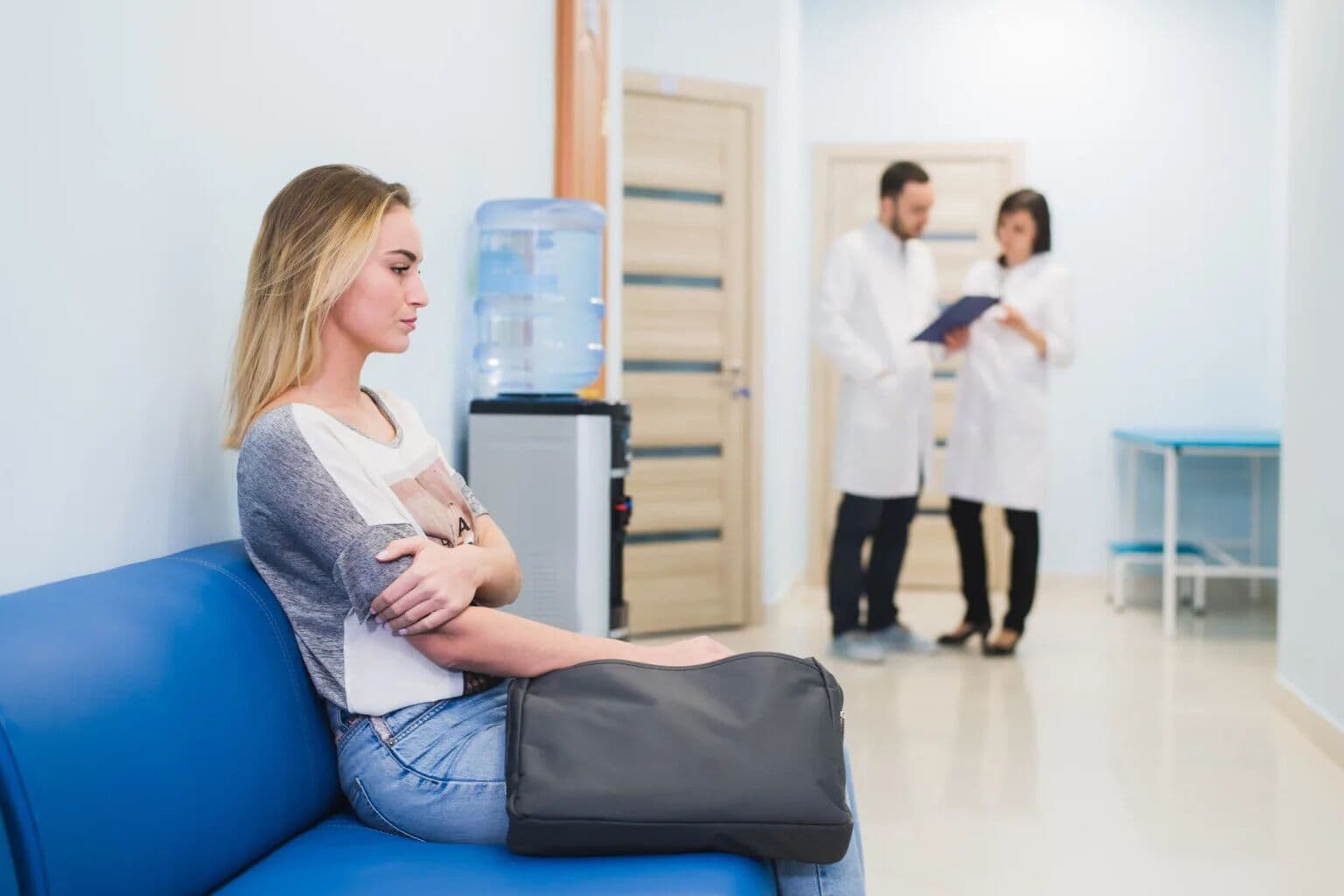 Paciente de braços cruzados espera sentada no sofá azul da sala de espera, enquanto um médico e uma médica conversam ao fundo, olhando uma prancheta
