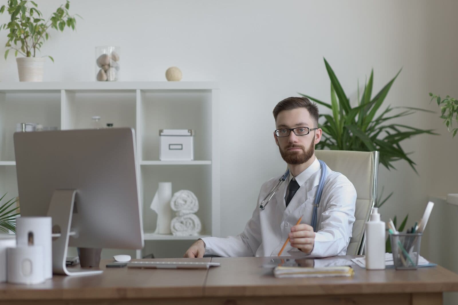 Médico de barba, óculos e jaleco, sentado à mesa do consultório com um lápis na mão, ao lado de um computador, com estante branca e plantas ao fundo