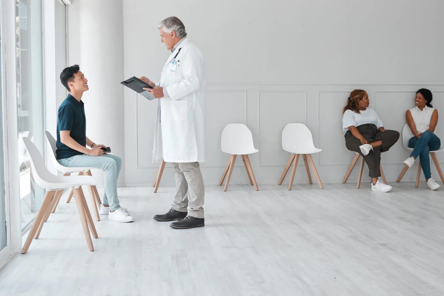 Médico de jaleco, com uma prancheta, conversa com um paciente sentado com o celular na mão, na sala de espera clara da clínica, enquanto duas pacientes aguardam nas cadeiras ao fundo