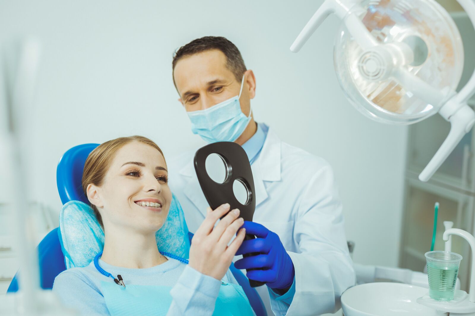 Paciente de aparelho ortodôntico sorri para o espelho de mão sentada na cadeira do consultório, ao lado do dentista de máscara, jaleco e luvas azuis