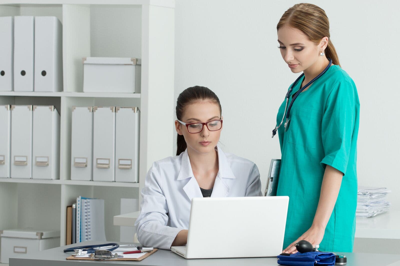 Médica de jaleco e óculos olha o notebook enquanto uma colega de uniforme verde, em pé ao lado, acompanha a mesma tela, num consultório com estante de pastas brancas
