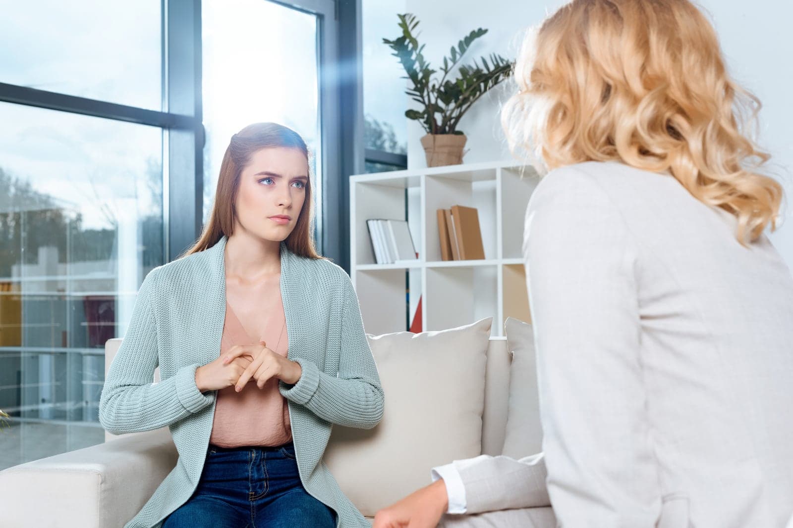 Mulher de jaleco branco, óculos e estetoscópio sorri sentada à mesa do consultório, com o celular nas mãos, ao lado do computador