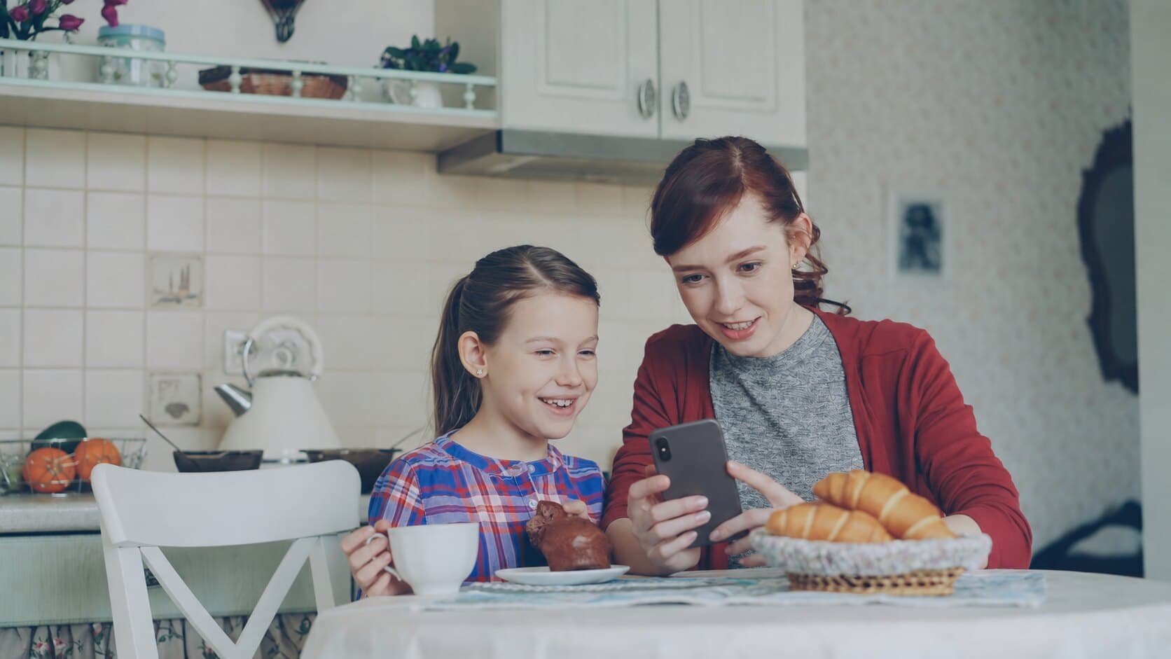 Mãe e filha na mesa do café da manhã, sorrindo ao ver a resposta no celular