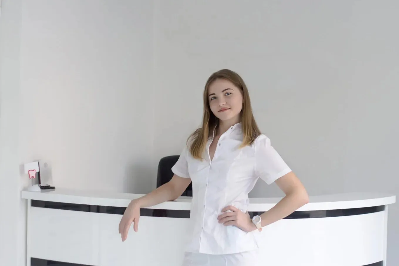 Recepcionista de uniforme branco sorri apoiada no balcão curvo da recepção de uma clínica