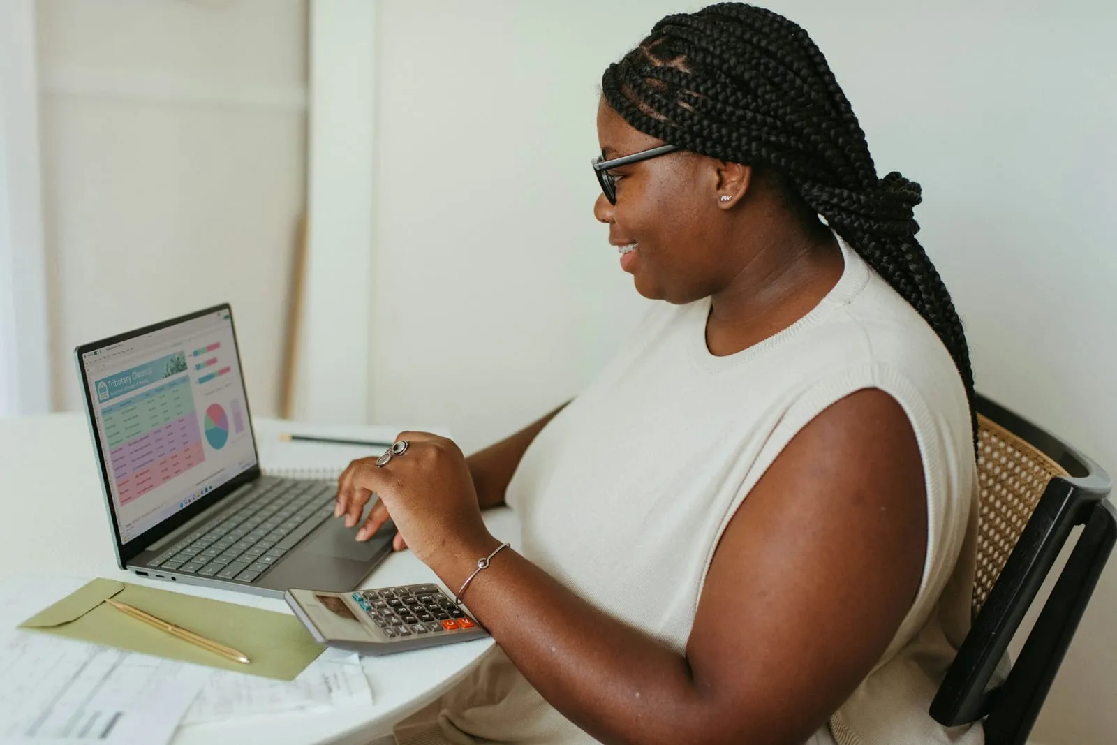 Mulher de tranças e óculos, sentada à mesa, digita numa calculadora ao lado do notebook aberto numa planilha com gráfico