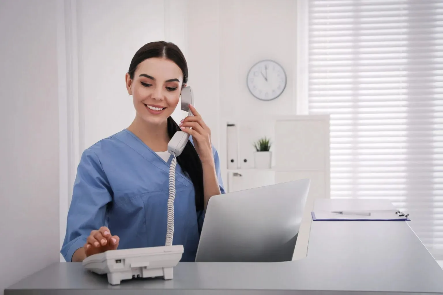 Recepcionista de uniforme azul sorri ao telefone no balcão da clínica, com um relógio na parede ao fundo