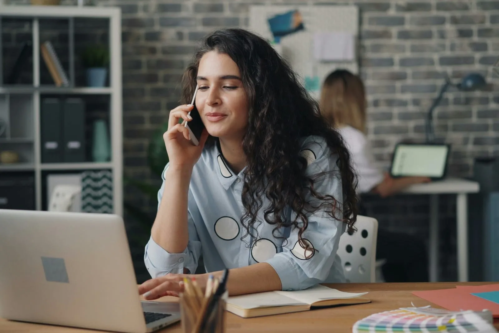 Jovem de cabelo cacheado fala ao celular sentada à mesa, com o notebook e um caderno aberto à frente, enquanto uma colega trabalha ao fundo
