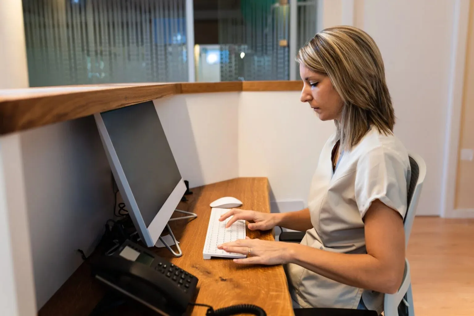 Recepcionista digita no computador atrás do balcão de madeira de uma clínica